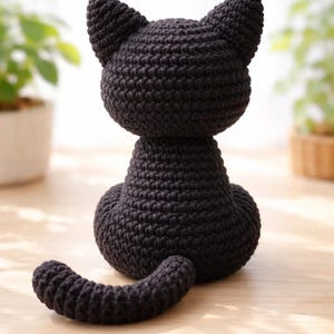 May include: A solid black crocheted cat figurine shown from the back. The cat has a rounded head with two pointed ears, a thick neck, a wide base, and a long, curved tail extending forward. The yarn texture is visible throughout the entire object.