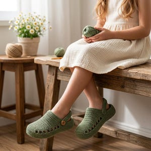 May include: A pair of sage green crochet clogs featuring a rounded toe, a perforated side design, and a small wooden button detail on the outer strap. The footwear is constructed with a textured yarn stitch and includes a back heel strap.