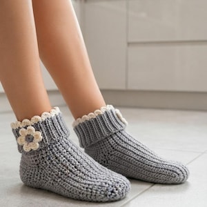 Può includere: Un paio di pantofole alla caviglia lavorate all'uncinetto in grigio con una trama a coste e un risvolto ripiegato. Ogni pantofola è decorata con un fiore all'uncinetto color crema sul lato. Il design presenta un bordo smerlato superiore.