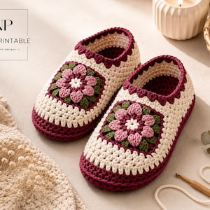 Può includere: Un paio di pantofole lavorate all'uncinetto a mano con una base color crema e un bordo bordeaux scuro. Ogni pantofola presenta un motivo quadrato sulla parte superiore, con un fiore rosa centrale e foglie verdi. Il design include un bordo smerlato.
