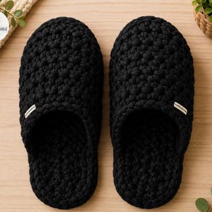 Op de afbeelding: Een paar zwarte slippers met een dikke, getextureerde gehaakte breiconstructie. Elke slipper heeft een klein, rechthoekig wit stoffen label aan de zijkant bij de opening. Het ontwerp is eenvoudig, met een ronde neus en een instapmodel.