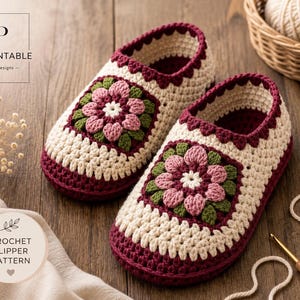 Op de afbeelding: Een paar handgehaakte slippers met een crèmekleurige basis, een donkerbordeauxrode rand en een centraal bloemmotief in roze, groene en witte tinten. De slippers hebben een geschulpte rand en een gestructureerd haakpatroon over het geheel.