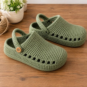 Könnte beinhalten: Ein Paar olivgrüne Häkel-Clogs mit strukturiertem Strickmuster, abgerundeter Zehenpartie und einem beweglichen Fersenriemen, der auf jeder Seite mit einem hellbraunen Holzknopf befestigt ist. Die Clogs haben rechteckige Ausschnitte an den Seiten.