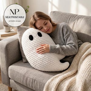 May include: A white ghost-shaped plush pillow with a textured, crocheted appearance and two solid black oval eyes. The pillow is soft and rounded, featuring a simple, minimalist design with a curved tail section extending to the right side.