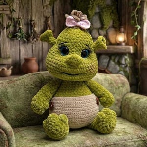 May include: A green crocheted plush toy with large dark eyes, long eyelashes, and trumpet-shaped ears. The figure has a small tuft of brown hair on top adorned with a light pink bow and wears a beige crocheted diaper secured with two brown buttons.