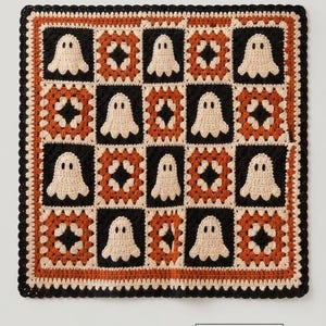 Pode incluir: Uma manta quadrada de crochê com um padrão repetitivo de quadrados pretos alternados com motivos de fantasmas na cor creme e quadrados de flores em laranja, preto e creme. A manta possui uma borda de crochê festonada na cor preta.