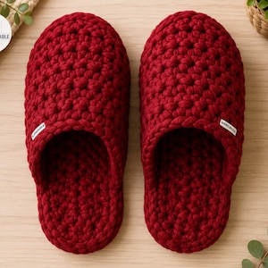 May include: A pair of deep red crochet slippers featuring a thick, textured yarn construction. Each slipper has a rounded toe, an open slip-on design, and a small white rectangular fabric tag attached to the side with black text.