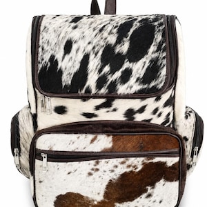 Puede incluir: Una mochila con estampado de piel de vaca, con manchas negras y blancas en la sección superior y manchas marrones y blancas en el bolsillo frontal. La bolsa incluye bordes de color marrón oscuro, cremalleras metálicas plateadas y bolsillos.