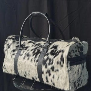 May include: A duffel bag with a spotted black and white pattern. The bag has black leather handles and straps, a zipper closure, and a silver-toned zipper pull. The bag is sitting on a black surface.
