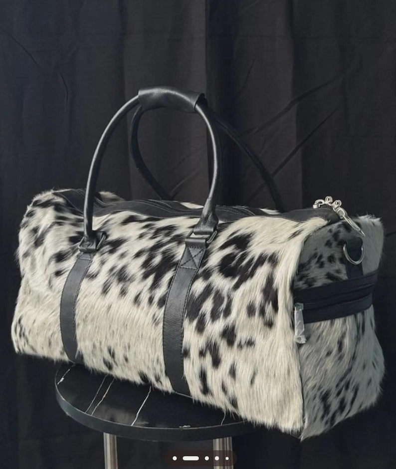 May include: A duffel bag with a spotted black and white pattern. The bag has black leather handles and straps, a zipper closure, and a silver-toned zipper pull. The bag is sitting on a black surface.