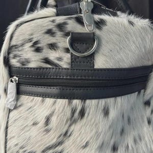 May include: A duffel bag with a spotted pattern in black and white. The bag features black leather accents, including a zipper, handle attachments, and a strap ring. The zipper pulls are silver. The bag's exterior has a soft, furry texture.
