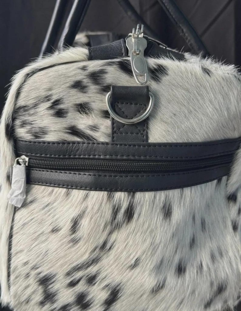 May include: A duffel bag with a spotted pattern in black and white. The bag features black leather accents, including a zipper, handle attachments, and a strap ring. The zipper pulls are silver. The bag's exterior has a soft, furry texture.