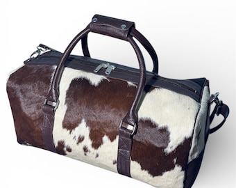 May include: A brown and white cowhide duffel bag with dark brown leather accents. The bag features a top handle, a zipper closure, and a detachable shoulder strap. The bag is designed for travel and has a classic, rustic aesthetic.