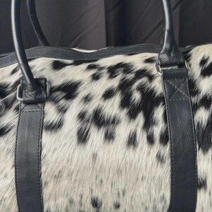 May include: A duffel bag with a spotted black and white pattern. The bag has black leather handles and straps. The bag is made of a soft, textured material.