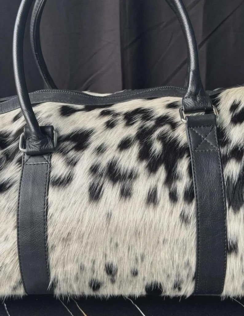 May include: A duffel bag with a spotted black and white pattern. The bag has black leather handles and straps. The bag is made of a soft, textured material.