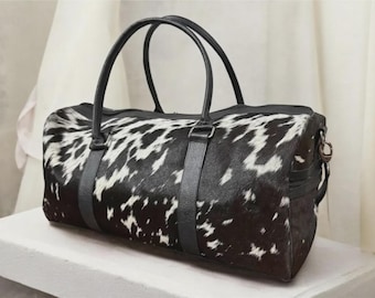 May include: A black and white cowhide duffel bag with black leather handles and trim. The bag has a cylindrical shape and a zipper closure. The bag is designed for travel and has a shoulder strap.