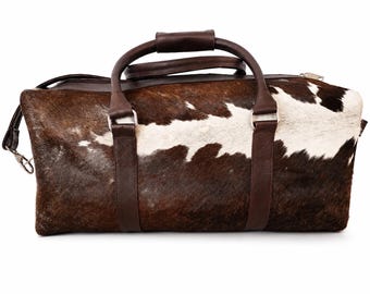 May include: A brown and white cowhide duffel bag with dark brown leather handles and straps. The bag has a zipper closure and a detachable shoulder strap. The bag is designed for travel and has a rectangular shape.