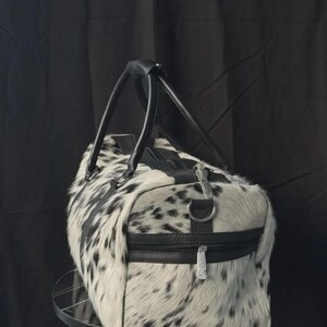 May include: A black and white cowhide duffel bag with black leather handles and trim. The bag has a zippered top and a side zipper pocket. The bag is cylindrical in shape and appears to be made of genuine cowhide.