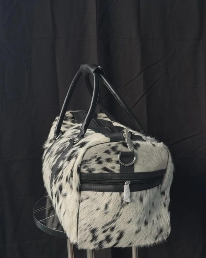 May include: A black and white cowhide duffel bag with black leather handles and trim. The bag has a zippered top and a side zipper pocket. The bag is cylindrical in shape and appears to be made of genuine cowhide.