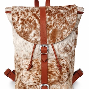 May include: A backpack featuring a brown and white cowhide pattern with smooth brown leather straps, a top handle, and two silver-toned metal buckles. The bag includes a front pocket with a buckle closure and adjustable side straps.
