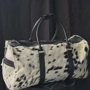 May include: A duffel bag with a spotted black and white pattern. The bag has black leather handles and trim. The bag is sitting on a small, round, black table.
