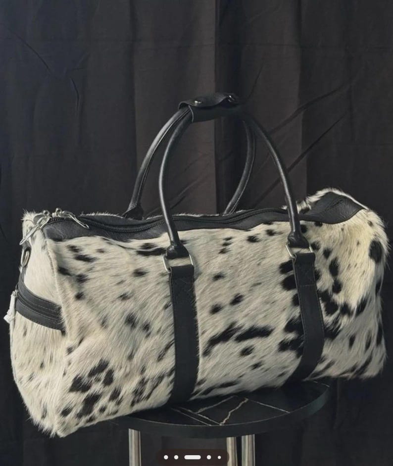 May include: A duffel bag with a spotted black and white pattern. The bag has black leather handles and trim. The bag is sitting on a small, round, black table.