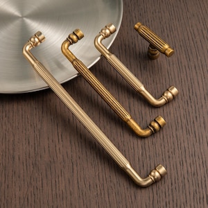 May include: A collection of antique brass cabinet pulls and a knob. The pulls are curved with a ribbed design, and the knob is a T-shape. The hardware is displayed on a dark wood surface.