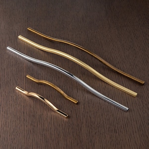 Op de afbeelding: Een assortiment gebogen metalen kastgrepen in verschillende afwerkingen, waaronder goud, zilver en brons. De grepen hebben een modern, minimalistisch ontwerp en liggen op een donkerhouten oppervlak.