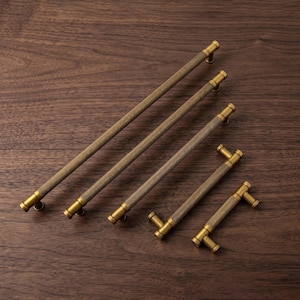 May include: A selection of brass cabinet pulls in different sizes, displayed on a dark wooden surface. The pulls feature a textured, ridged central section and rounded ends. The hardware has a warm, antique brass finish.