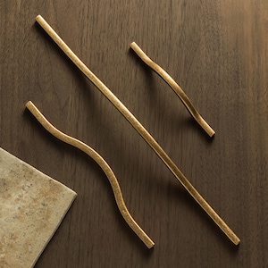 Pode incluir: Três puxadores dourados para armários em vários designs. Um é reto e dois são curvos. O hardware é feito de metal e tem um acabamento texturizado. Os puxadores são exibidos em uma superfície de madeira escura.