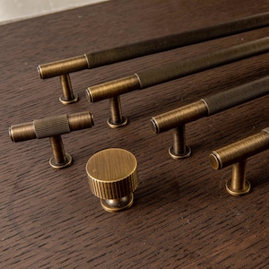 Puede incluir: Un surtido de herrajes de gabinete de latón antiguo, que incluye varios tiradores en forma de T y un pomo redondo. Los herrajes presentan un diseño acanalado en las manijas y perillas, sobre una superficie de madera oscura.