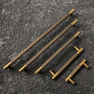 May include: Four antique brass cabinet pulls in varying sizes. The pulls have a textured, ridged center section and are mounted with brass hardware. The hardware is a T-shape or a curved bar. The cabinet pulls are arranged diagonally on a dark, textured surface.