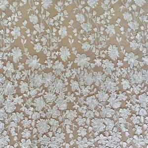 K&ouml;nnte beinhalten: Wei&szlig;er floraler Spitzenstoff mit einem detaillierten Muster aus verschiedenen Blumenarten und Bl&auml;ttern. Die Spitze ist dicht bestickt und erzeugt eine strukturierte Oberfl&auml;che. Der Hintergrund ist hellbeige und bildet einen Kontrast zur wei&szlig;en Spitze.