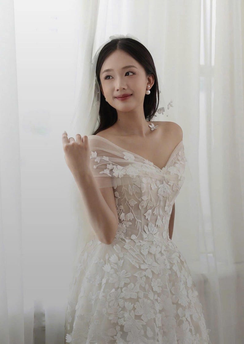 K&ouml;nnte beinhalten: Wei&szlig;es schulterfreies Brautkleid mit floralen Spitzendetails. Das Kleid hat einen Sweetheart-Ausschnitt und einen weiten Rock. Ein durchsichtiger Schleier und Perlenohrringe vervollst&auml;ndigen den Braut-Look.