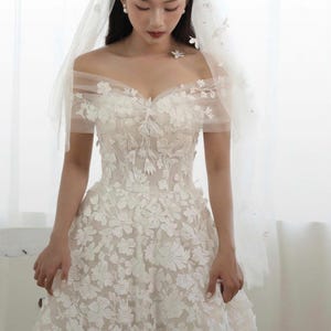 K&ouml;nnte beinhalten: Wei&szlig;es Hochzeitskleid mit floraler Stickerei und schulterfreiem Ausschnitt. Das Kleid hat einen weiten Rock und wird mit einem transparenten Schleier kombiniert, der mit zarten floralen Akzenten verziert ist. Die Braut tr&auml;gt Perlenohrringe.