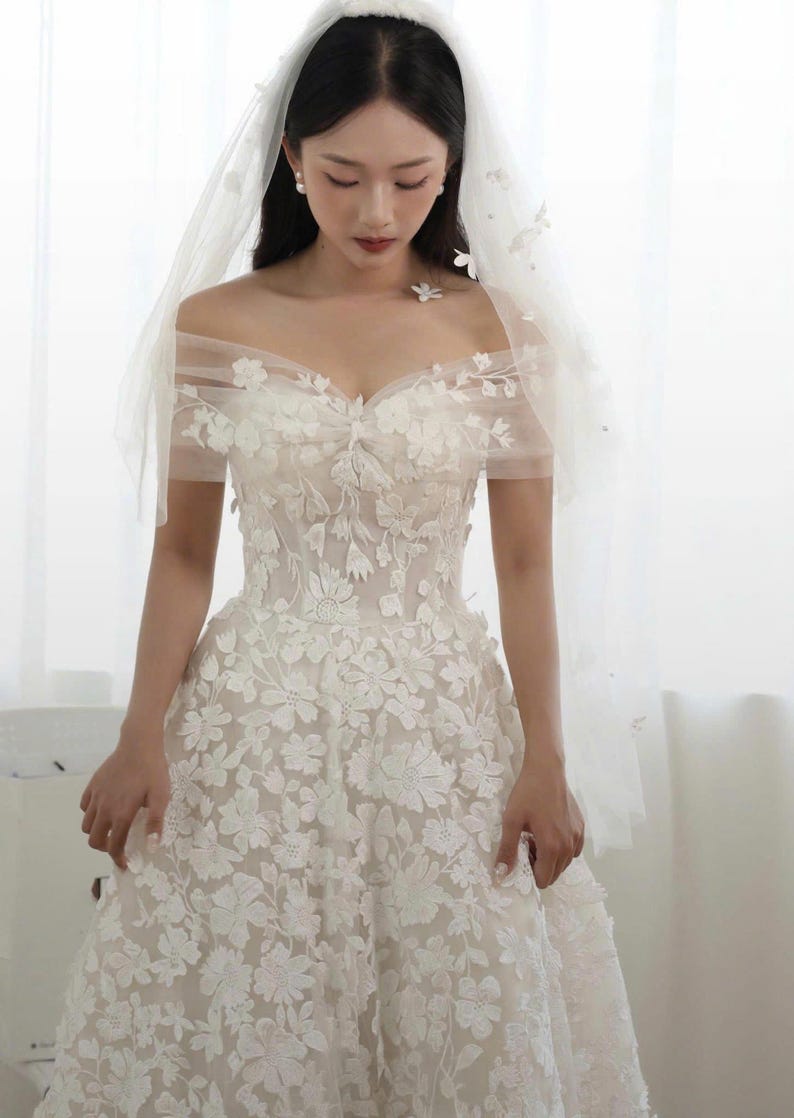 K&ouml;nnte beinhalten: Wei&szlig;es Hochzeitskleid mit floraler Stickerei und schulterfreiem Ausschnitt. Das Kleid hat einen weiten Rock und wird mit einem transparenten Schleier kombiniert, der mit zarten floralen Akzenten verziert ist. Die Braut tr&auml;gt Perlenohrringe.