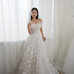 K&ouml;nnte beinhalten: Ein wei&szlig;es schulterfreies Hochzeitskleid mit floralen Applikationen. Das Kleid hat ein eng anliegendes Oberteil und einen flie&szlig;enden Rock. Das Kleid wird vor einem Fenster mit wei&szlig;en Vorh&auml;ngen pr&auml;sentiert.