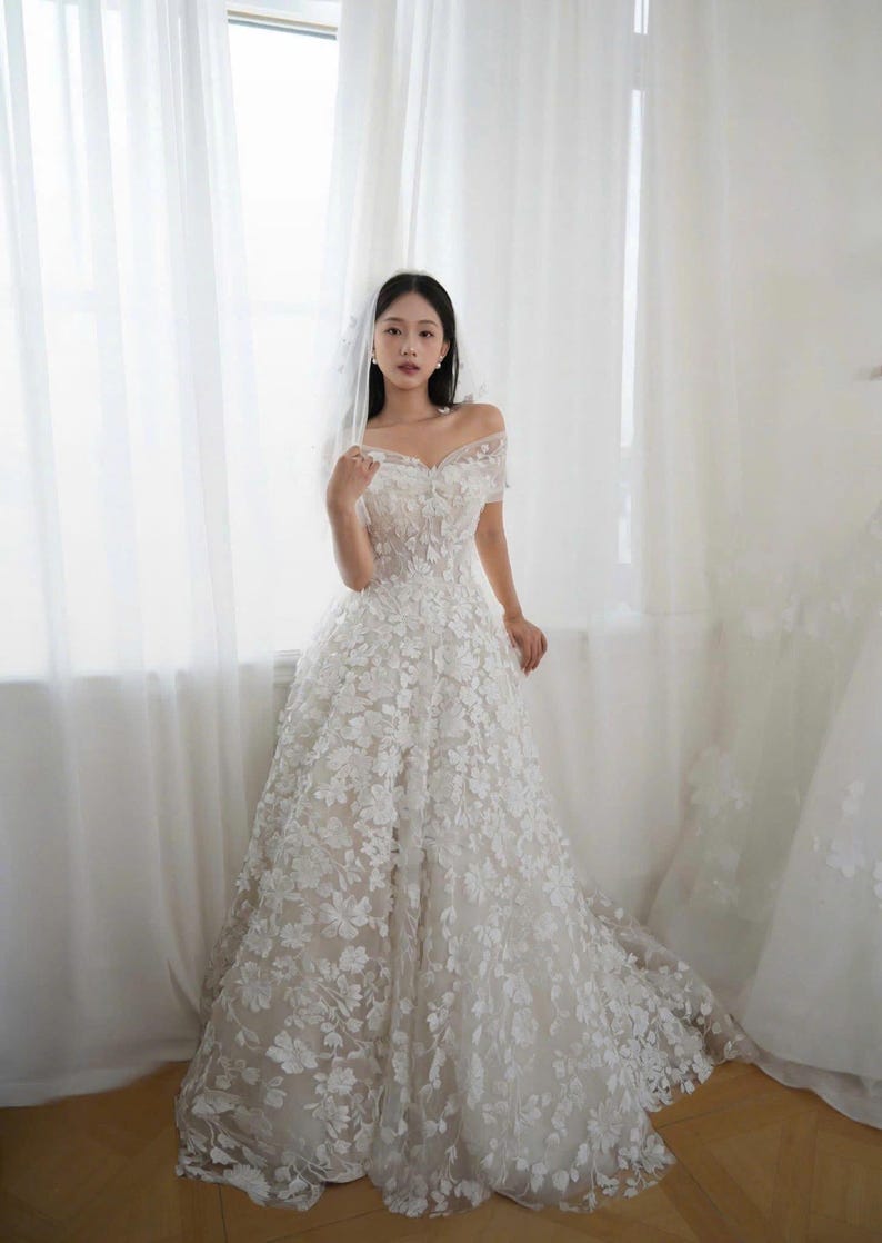 K&ouml;nnte beinhalten: Ein wei&szlig;es schulterfreies Hochzeitskleid mit floralen Applikationen. Das Kleid hat ein eng anliegendes Oberteil und einen flie&szlig;enden Rock. Das Kleid wird vor einem Fenster mit wei&szlig;en Vorh&auml;ngen pr&auml;sentiert.