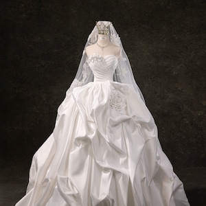 Könnte beinhalten: Weißes, trägerloses Brautkleid aus Satin mit weitem Rock und Rosendetail. Das Kleid wird auf einer Schaufensterpuppe präsentiert und mit Schleier, Krone und Halskette kombiniert. Der Hintergrund ist dunkel und strukturiert.