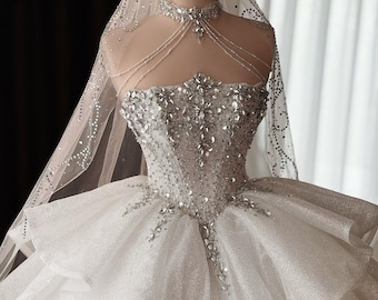 Può includere: Abito da sposa bianco con corpetto scintillante e gonna a strati, esposto su un manichino. L'abito presenta intricate perline argentate e un velo trasparente ornato di cristalli. Una corona d'oro è posta sulla testa del manichino.