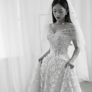 K&ouml;nnte beinhalten: Wei&szlig;es Hochzeitskleid mit floralen Applikationen und einem Sweetheart-Ausschnitt. Die schulterfreien &Auml;rmel sind mit zarten floralen Details verziert. Ein durchsichtiger Schleier ist &uuml;ber dem Kopf drapiert und erg&auml;nzt den Braut-Look.