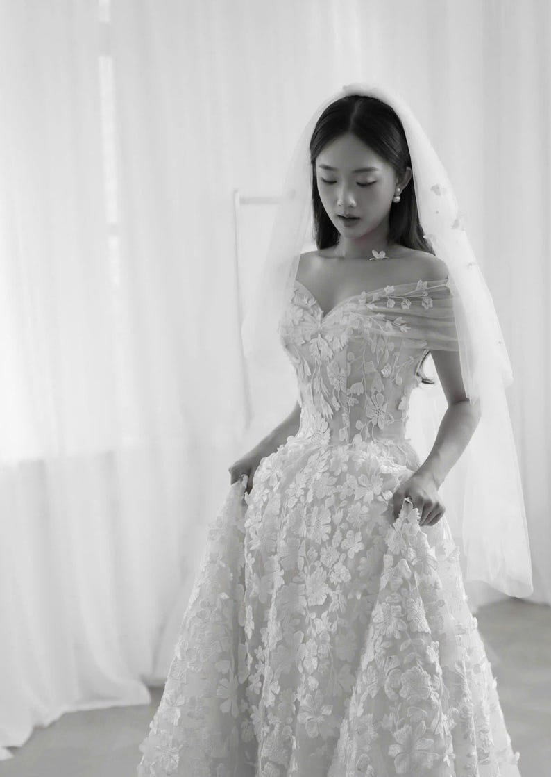 K&ouml;nnte beinhalten: Wei&szlig;es Hochzeitskleid mit floralen Applikationen und einem Sweetheart-Ausschnitt. Die schulterfreien &Auml;rmel sind mit zarten floralen Details verziert. Ein durchsichtiger Schleier ist &uuml;ber dem Kopf drapiert und erg&auml;nzt den Braut-Look.