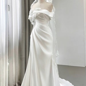 May include: A white satin wedding dress with an off-the-shoulder neckline and a flowing skirt. The dress features delicate floral embellishments and is paired with a sheer veil with lace trim. The gown is displayed on a mannequin.