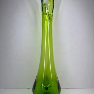 May include: A tall, lime green glass vase with a slender, elongated neck and a flared, irregular rim that resembles a splash. The base of the vase is rounded and supported by three small, integrated glass feet, creating a tripod-like structure.