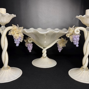 May include: A set of three decorative glass pieces: two candelabras and a centerpiece bowl. The candelabras have twisted stems, grape clusters, and leaf accents. The bowl has a ruffled edge and a twisted base. All pieces are off-white.