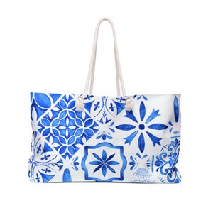 May include: A large tote bag featuring a white base with a repeating blue watercolor tile pattern. The bag has two white rope handles attached with silver-toned metal grommets. The design includes various geometric and floral motifs in shades of blue.