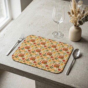 May include: A rectangular placemat featuring a repeating grid pattern of autumn-themed squares in shades of orange, green, yellow, and brown, decorated with small illustrations of pumpkins, leaves, and acorns, accompanied by a fork, spoon, and glass.