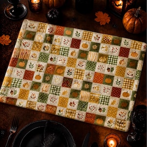 May include: A rectangular quilted placemat featuring a patchwork grid of squares in autumn colors including orange, green, yellow, and brown. The fabric squares are decorated with printed motifs of pumpkins, acorns, leaves, and plaid patterns.