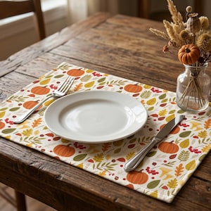 May include: A rectangular placemat featuring a repeating pattern of orange pumpkins, red berries, and multicolored autumn leaves on a cream background. A white ceramic plate, a silver fork, and a silver knife are placed on the placemat.