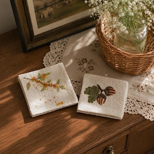 May include: Two square, off-white embroidered linen coasters. One features a branch with yellow flowers and green leaves, the other has an acorn and oak leaf design. The coasters are on a wooden surface, with a framed picture and a basket in the background.